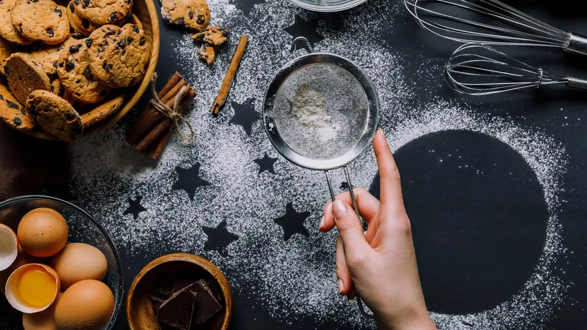 Ingrediencie a ruka pri práci – okraje s vajíčkami, čokoládou a škoricou, košík s cookies, metlička a ruka, ktorá preosieva múku: Príprava dezertov – kvalitné ingrediencie a ručná práca. Vajíčka, čokoláda, škorica, košík s cookies a ruka preosievajúca múku sitkom.